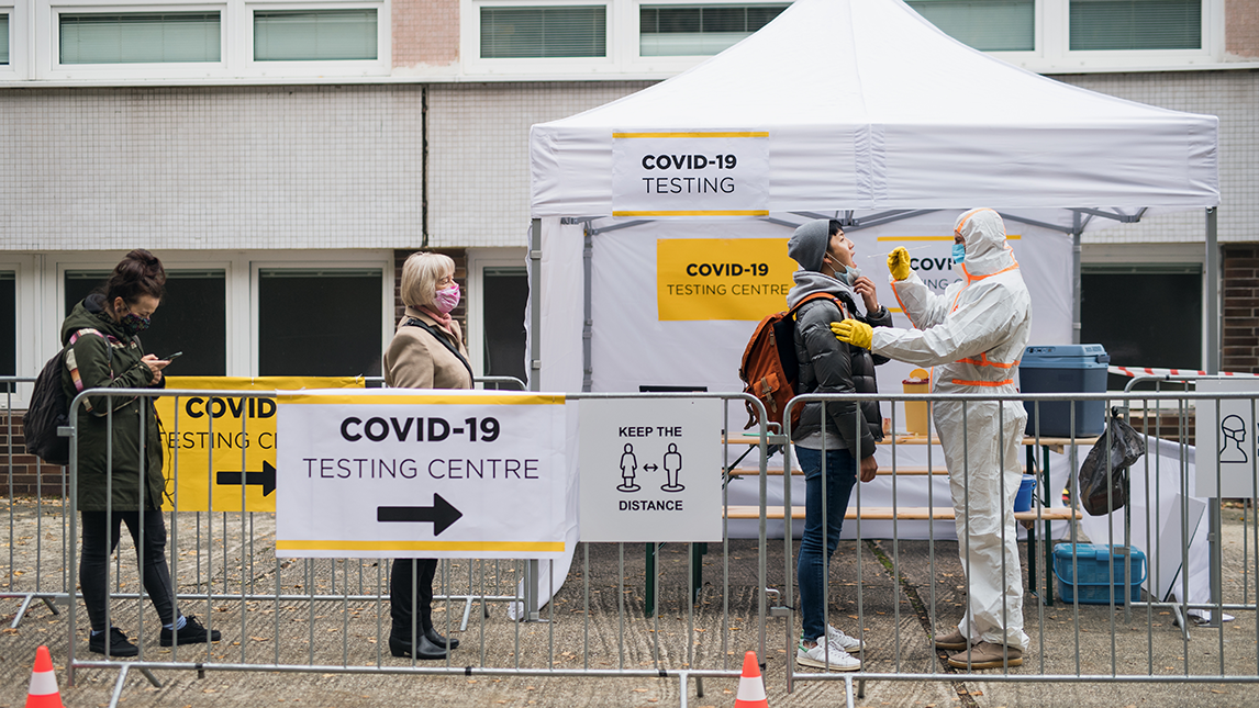 Person being tested for COVID-19 at an outdoor testing centre by someone in PPE while two people wait in line.