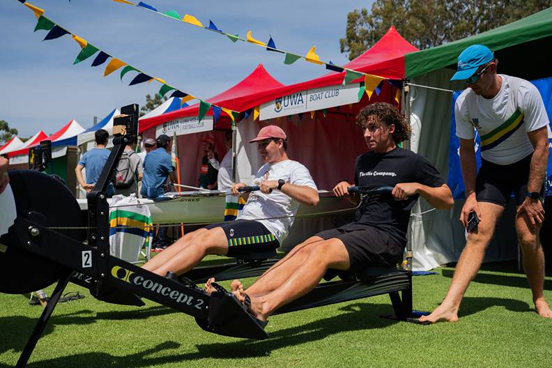 Two male students sitting on rowing machines competing outdoors at O-Week, one male student standing behind as an onlooker. In the background, O-Week marquees are set up, including 'UWA BOAT CLUB'