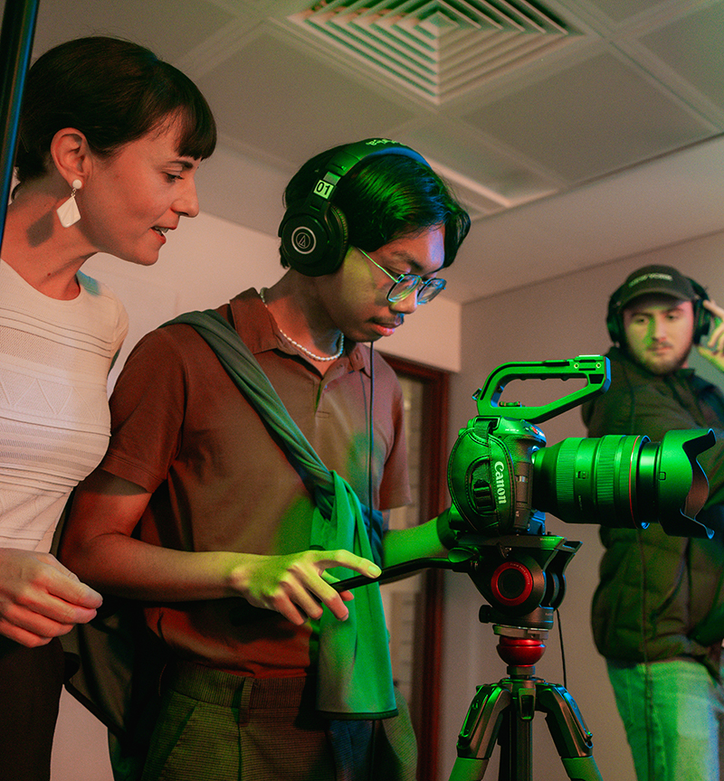 A student wearing headphones looks into a camera while an academic stands next to them, providing instruction. In the background, another student is wearing headphones and holding a boom pole.