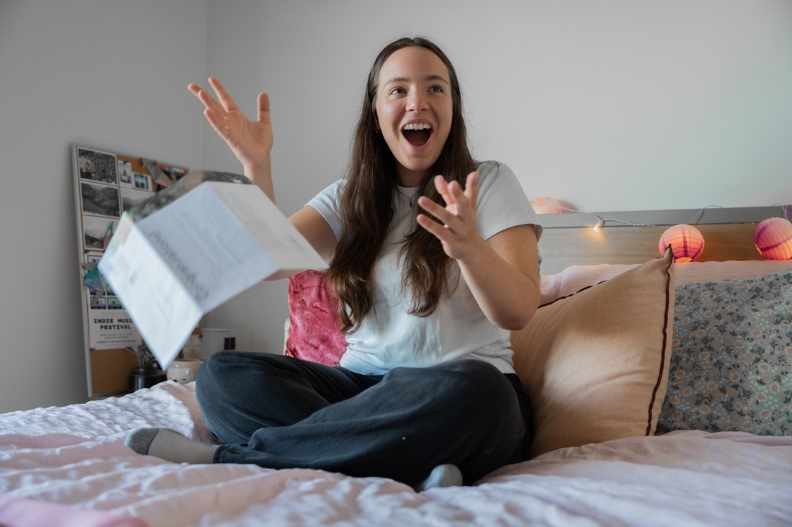 Student celebrating receiving acceptance letter, sitting on top of the bed in bedroom